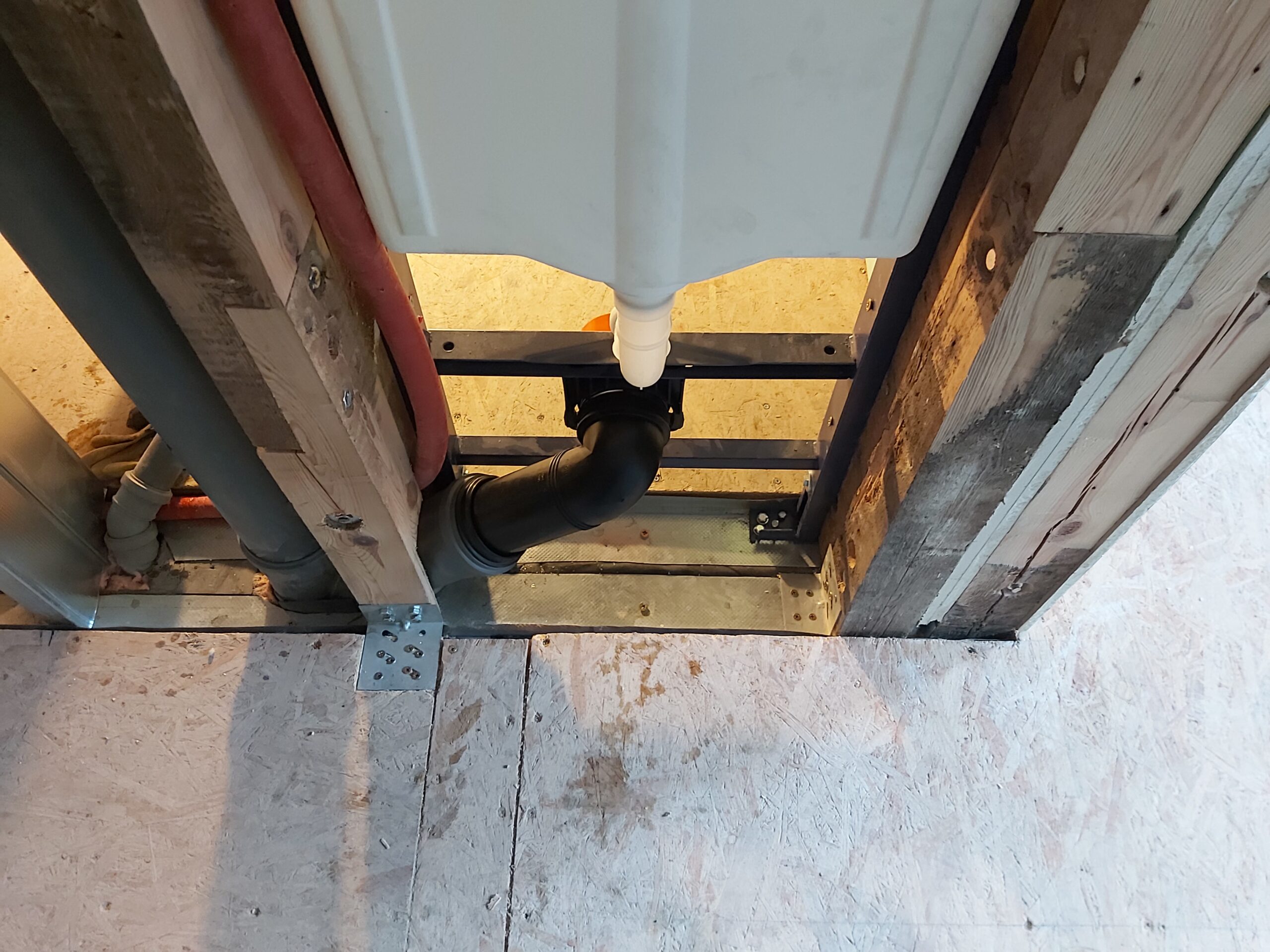 Dolna część podtynkowego stelaża WC z widocznym odpływem kanalizacyjnym, mocowaniem do podłogi oraz elementami konstrukcji drewniano-metalowej przed zabudową.