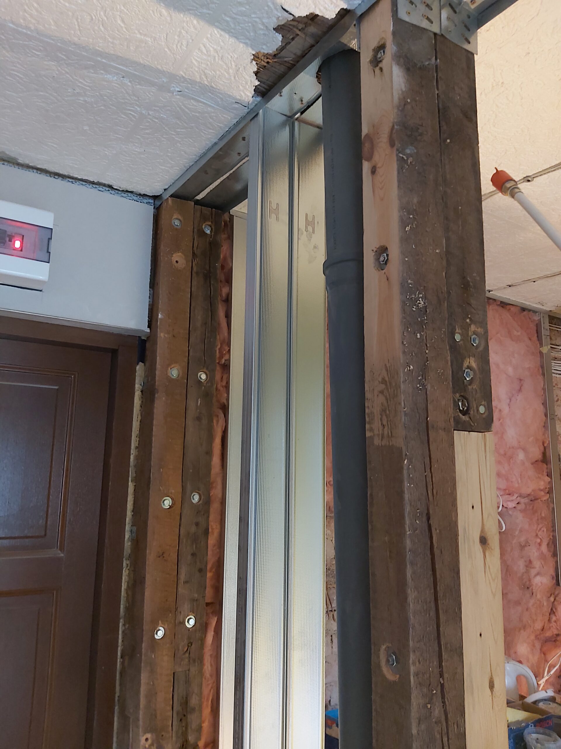 Fragment konstrukcji ściany szkieletowej przy drzwiach z widocznym pionem kanalizacyjnym i profilami stalowymi