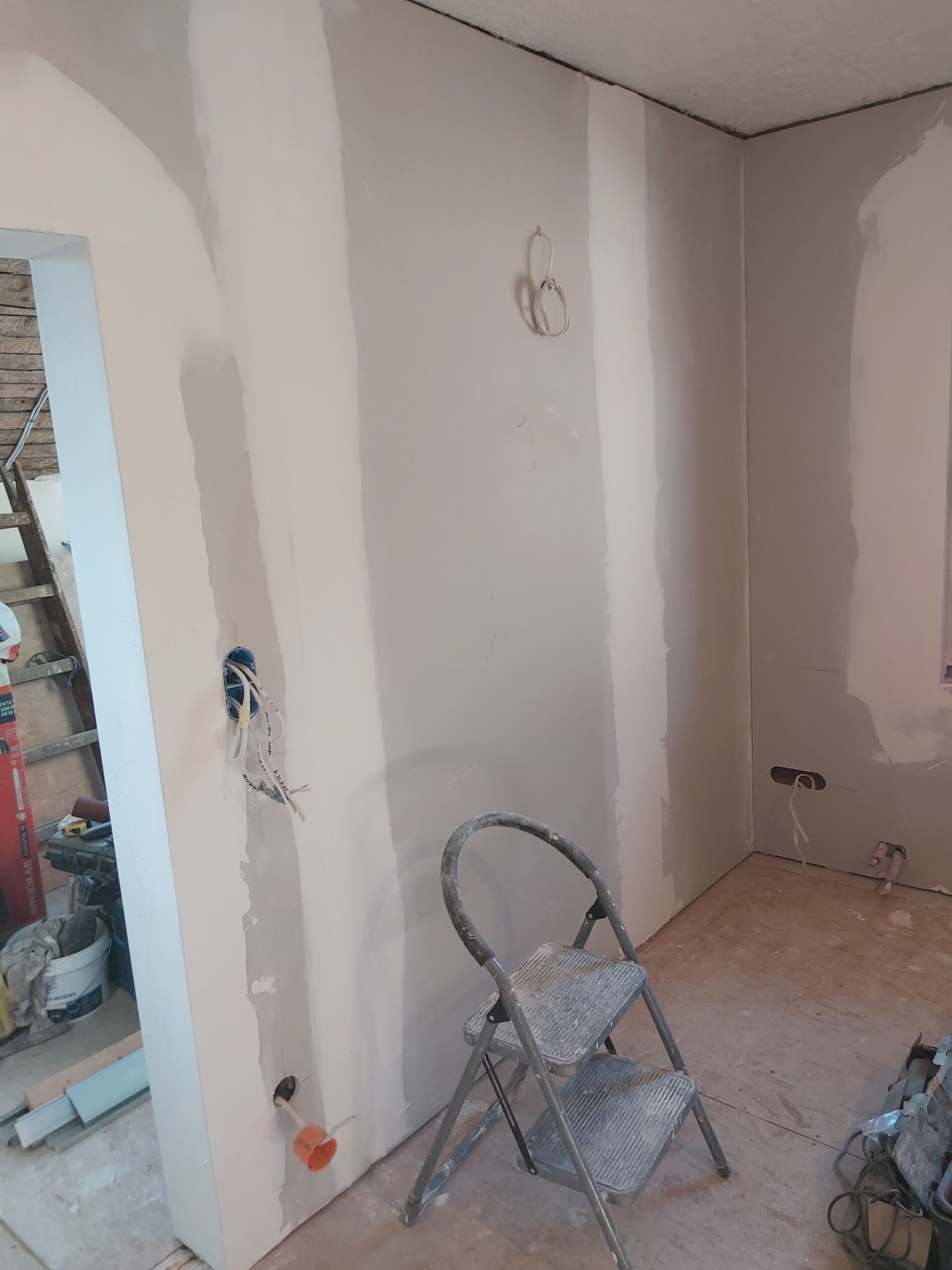 Wnętrze pomieszczenia po szpachlowaniu ścian z płyt gipsowo-kartonowych, z widocznymi pasami gładzi, wyprowadzonymi przewodami elektrycznymi, otworem drzwiowym oraz drabiną roboczą ustawioną przy ścianie.