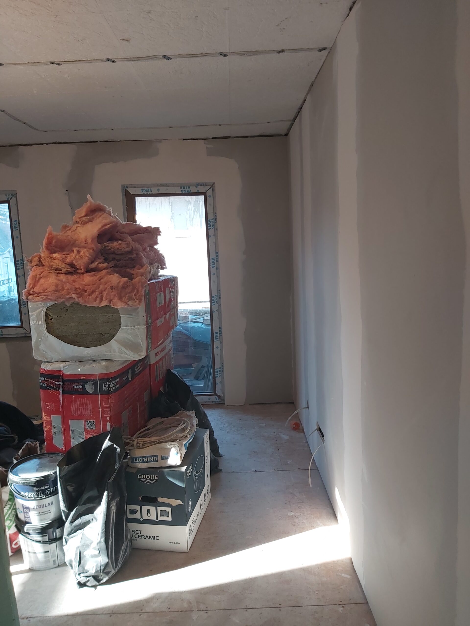 Pomieszczenie w trakcie wykańczania, z wygładzonymi ścianami z płyt gipsowo-kartonowych po pierwszym szpachlowaniu, widocznym oknem, surowym stropem oraz materiałami budowlanymi i izolacją ułożonymi na podłodze.