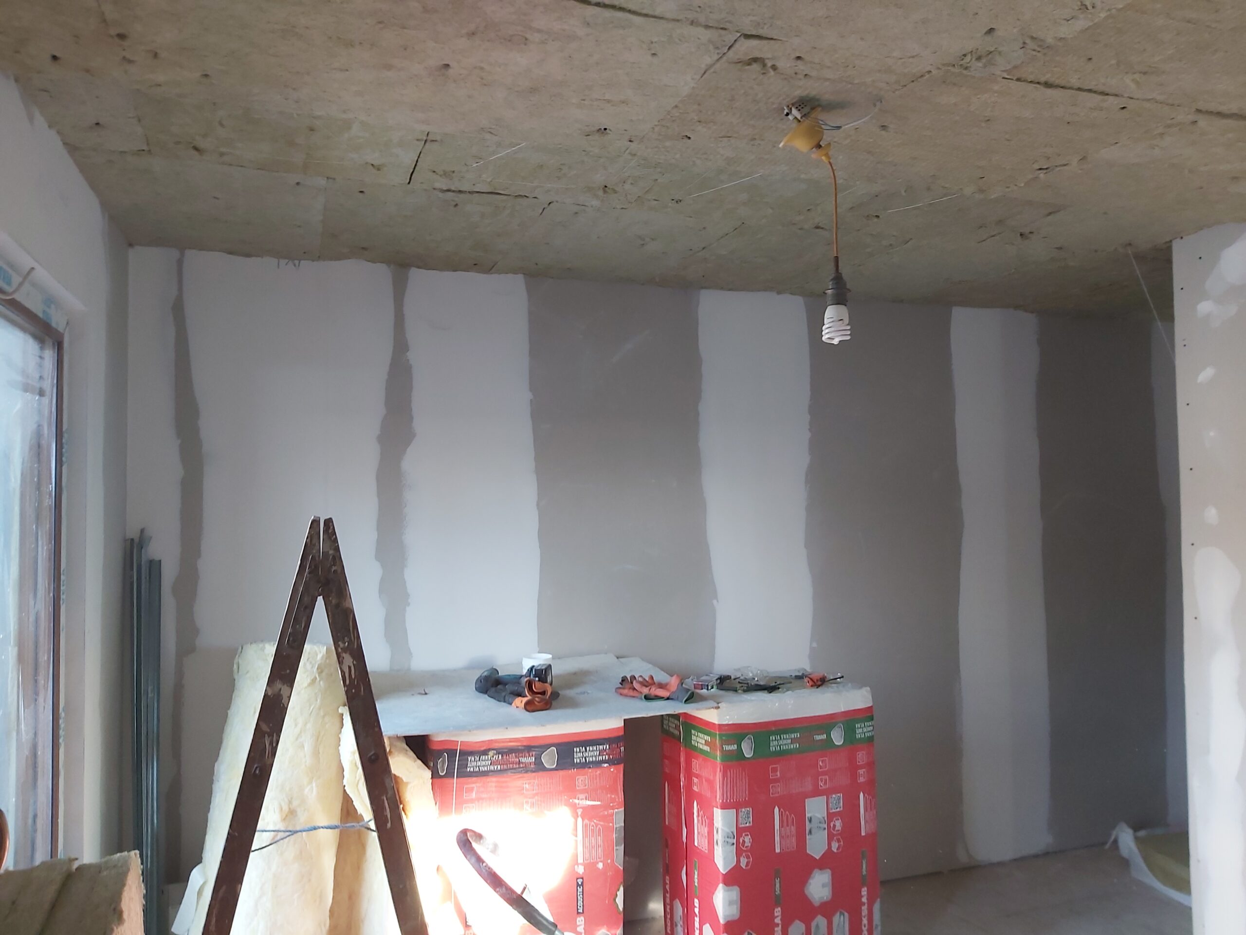 Ściana z płyt gipsowo-kartonowych w trakcie wykańczania, z pionowymi pasami masy szpachlowej na łączeniach, surowym stropem betonowym oraz materiałami budowlanymi ustawionymi przy ścianie.