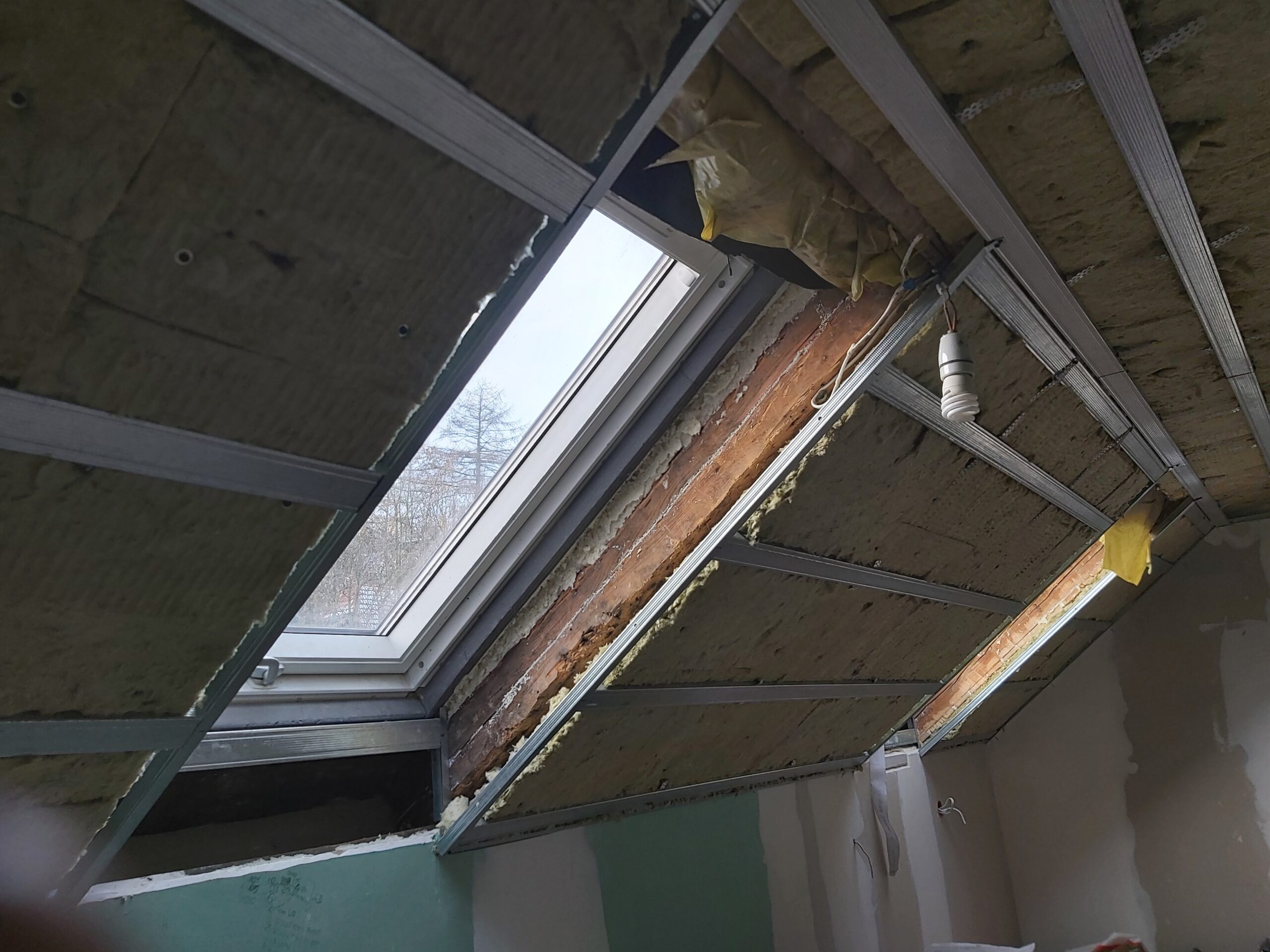 Okno dachowe podczas prac remontowych na poddaszu z widoczną konstrukcją metalową i izolacją