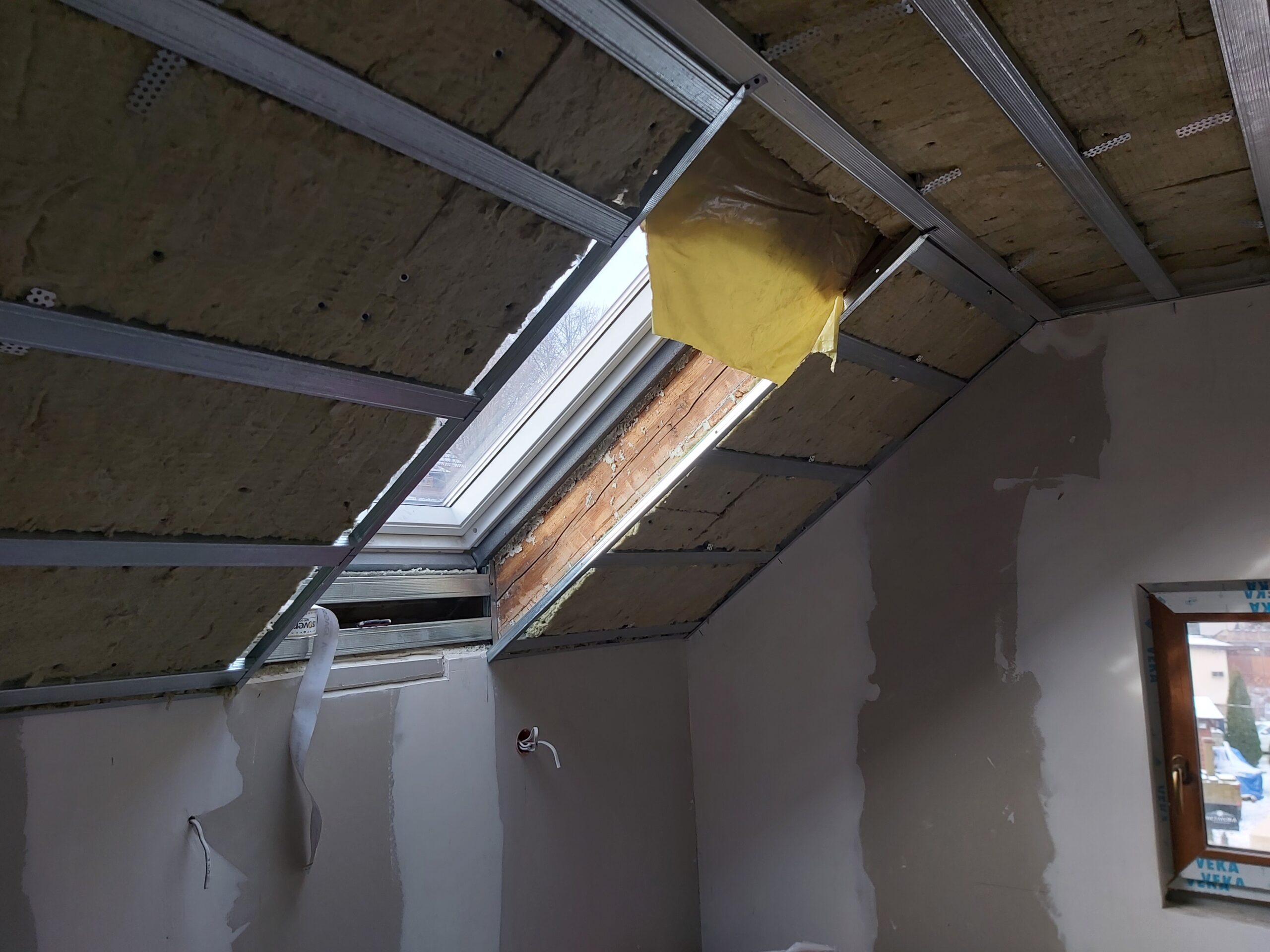 Okno dachowe w trakcie montażu zabudowy poddasza, widoczna konstrukcja metalowa i izolacja