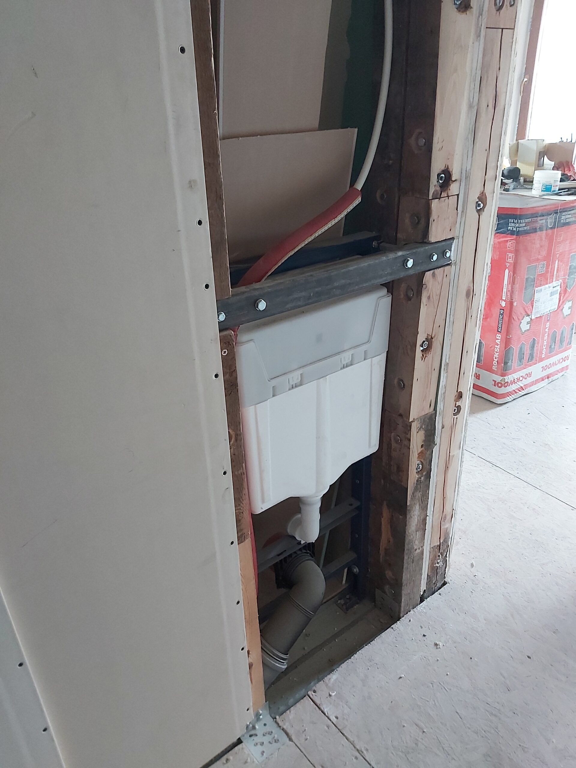 Podtynkowy stelaż WC zamontowany w ścianie szkieletowej, z widoczną konstrukcją drewnianą, instalacją kanalizacyjną i częściowo zamontowaną płytą gipsowo-kartonową podczas prac remontowych.
