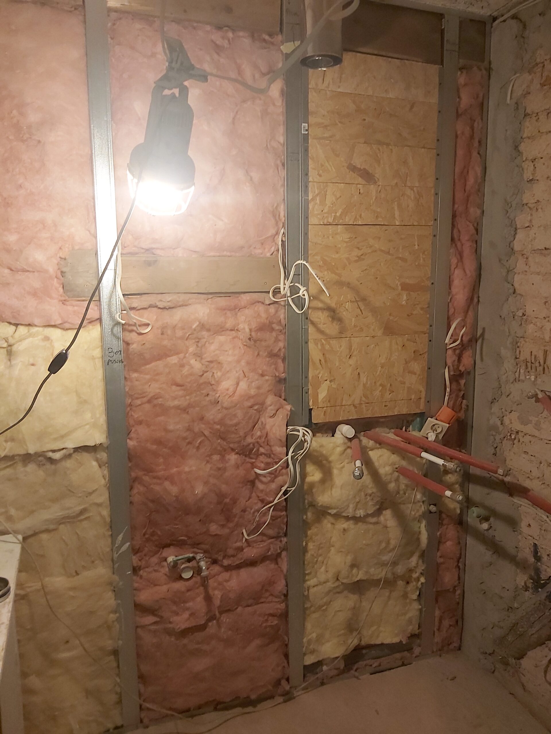 Ściana w trakcie remontu z metalowym stelażem, wełną izolacyjną w kolorze różowym i żółtym, widocznymi rurami instalacyjnymi, przewodami elektrycznymi oraz lampą roboczą.