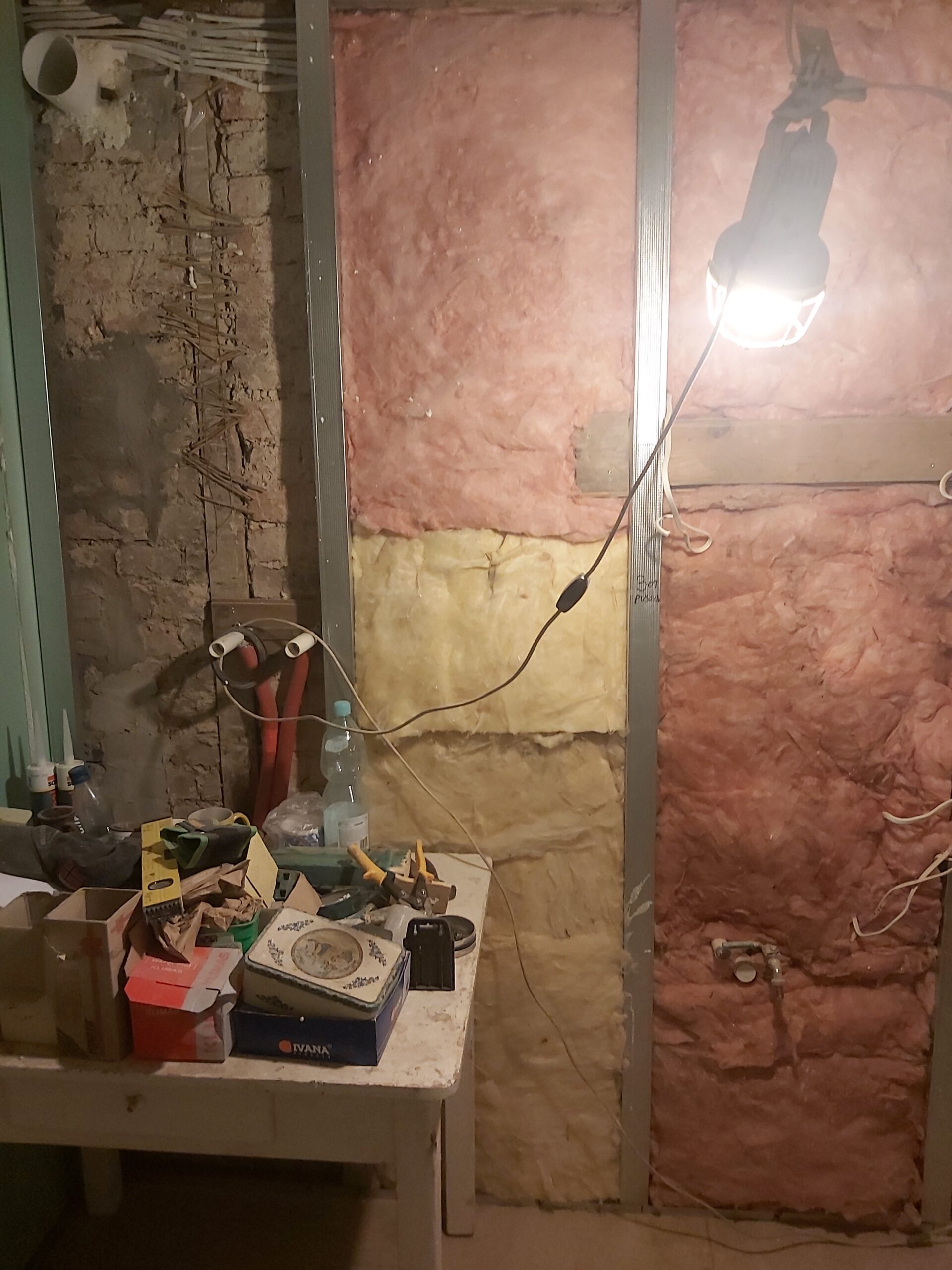 Ściana w trakcie remontu z widoczną wełną izolacyjną w kolorze różowym i żółtym, metalowym stelażem, przewodami elektrycznymi oraz tymczasowym oświetleniem roboczym.