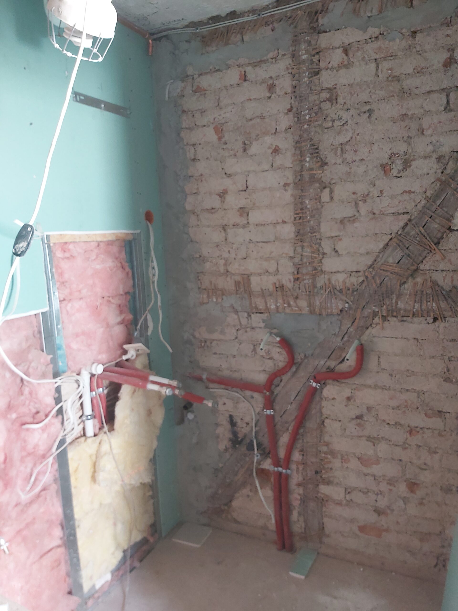 Odsłonięta ceglana ściana z widoczną instalacją hydrauliczną i elektryczną podczas remontu pomieszczenia, z poprowadzonymi czerwonymi rurami, przewodami oraz fragmentami izolacji.