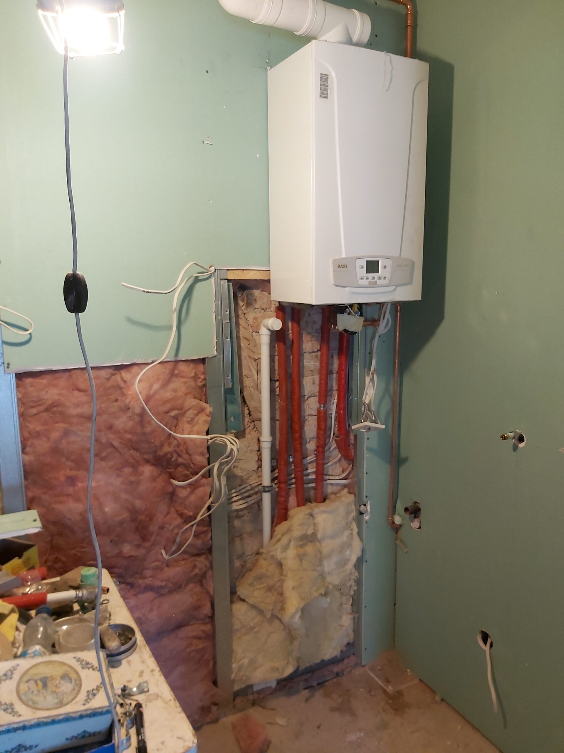 Kocioł gazowy zamontowany na ścianie w trakcie remontu, z odsłoniętą instalacją hydrauliczną, przewodami elektrycznymi, wełną mineralną oraz konstrukcją pod zabudowę z płyt gipsowo-kartonowych.