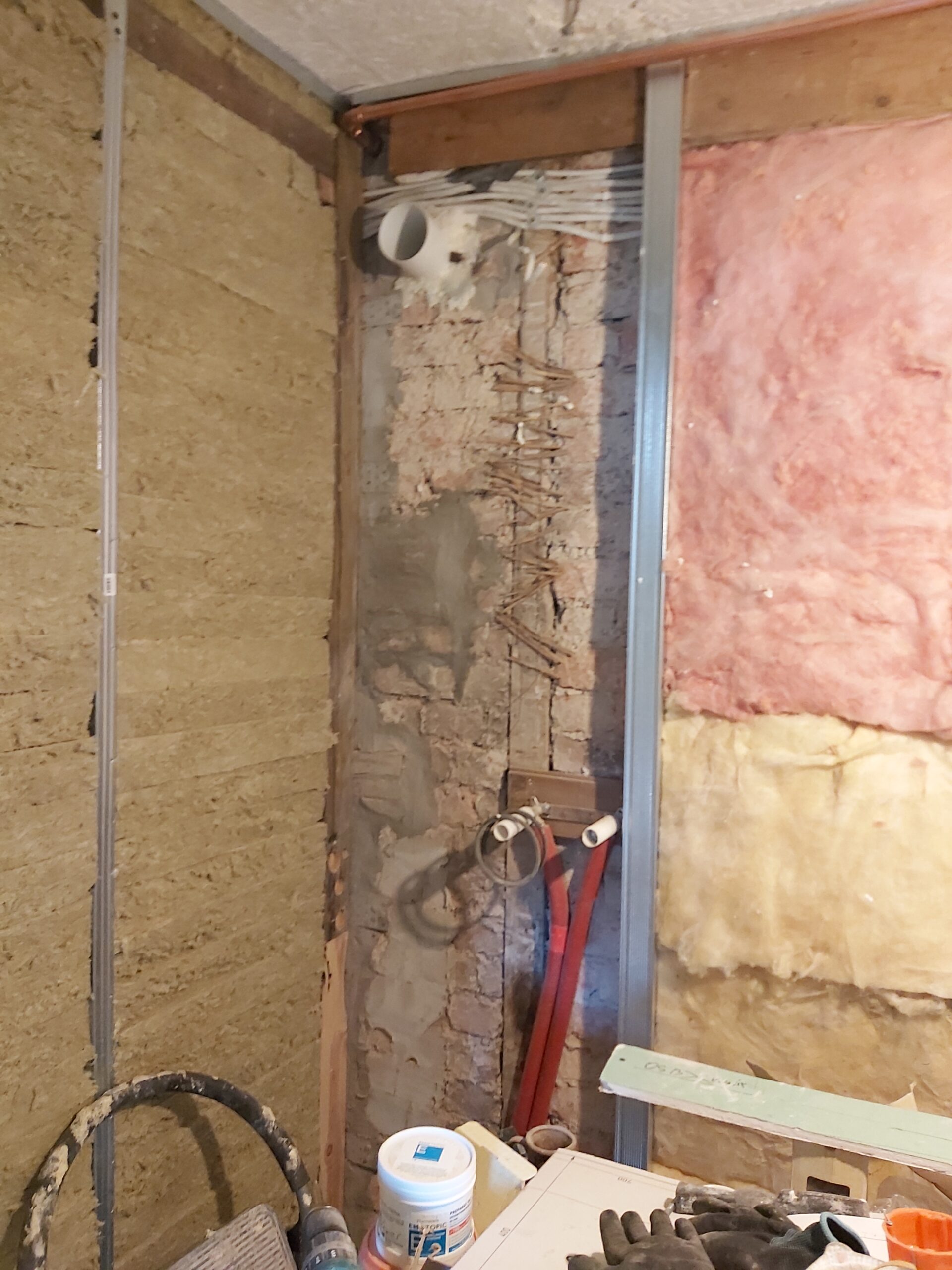 Fragment ściany w trakcie remontu z widoczną instalacją elektryczną i hydrauliczną, wełną mineralną oraz profilami stalowymi pod zabudowę z płyt gipsowo-kartonowych.
