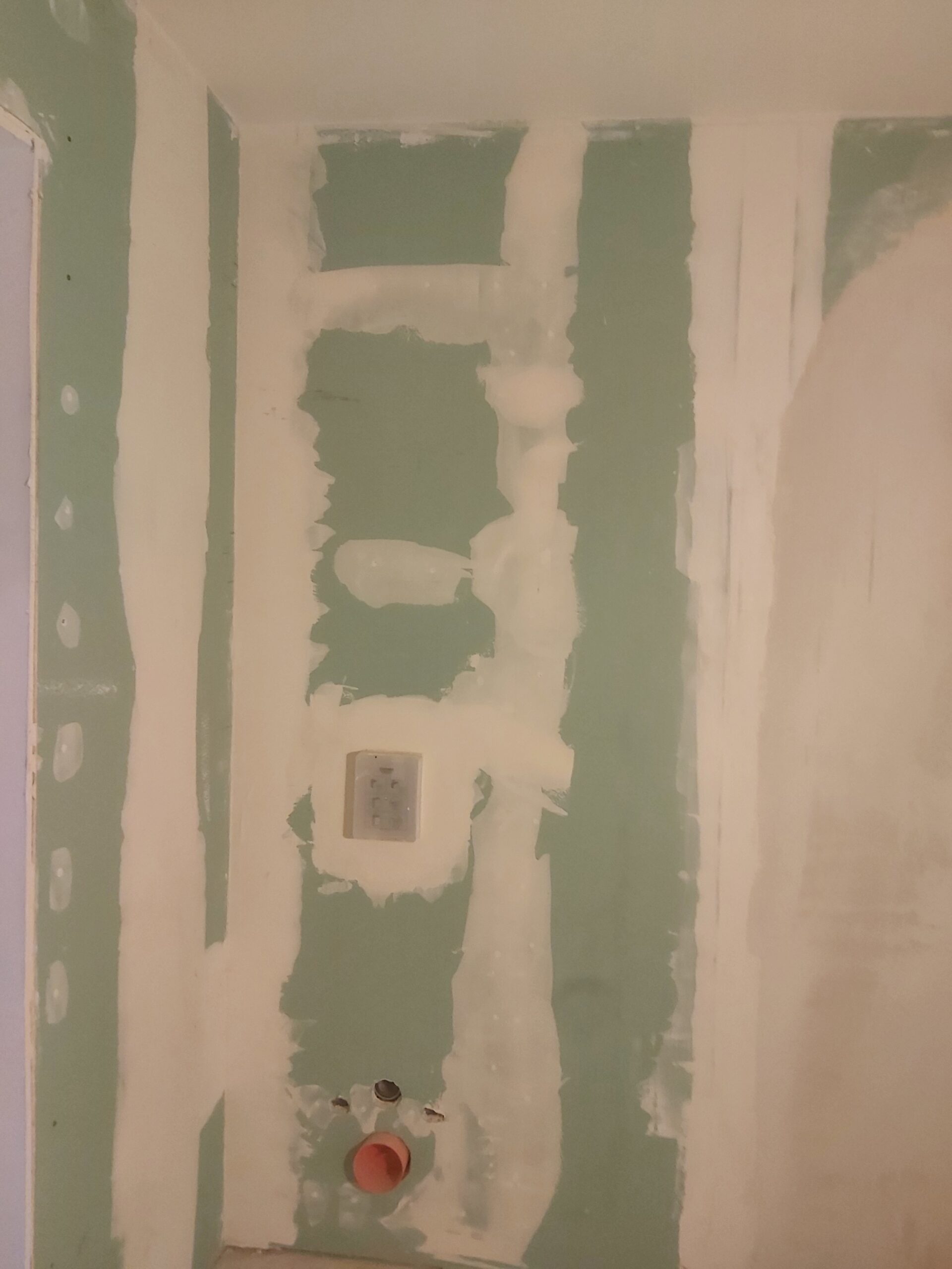 Zielona ściana z płyt gipsowo-kartonowych w trakcie szpachlowania, z widocznymi pasami masy na łączeniach płyt oraz przygotowanymi wyjściami instalacji wodno-kanalizacyjnej i puszką elektryczną przed dalszym wykończeniem.