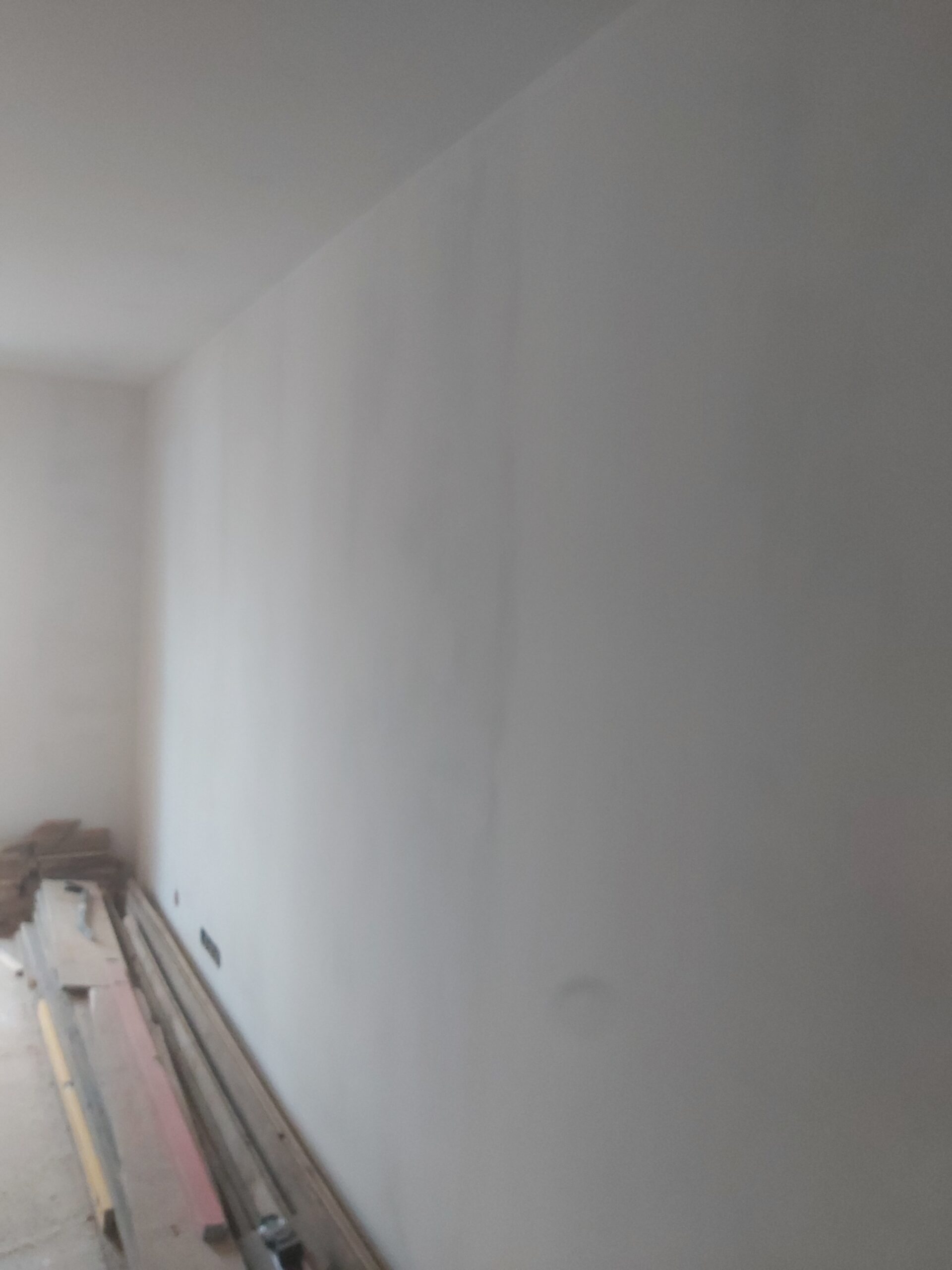 Długa, biała ściana po szpachlowaniu i malowaniu, z widocznymi smugami po pracach wykończeniowych oraz materiałami budowlanymi ułożonymi przy podłodze.