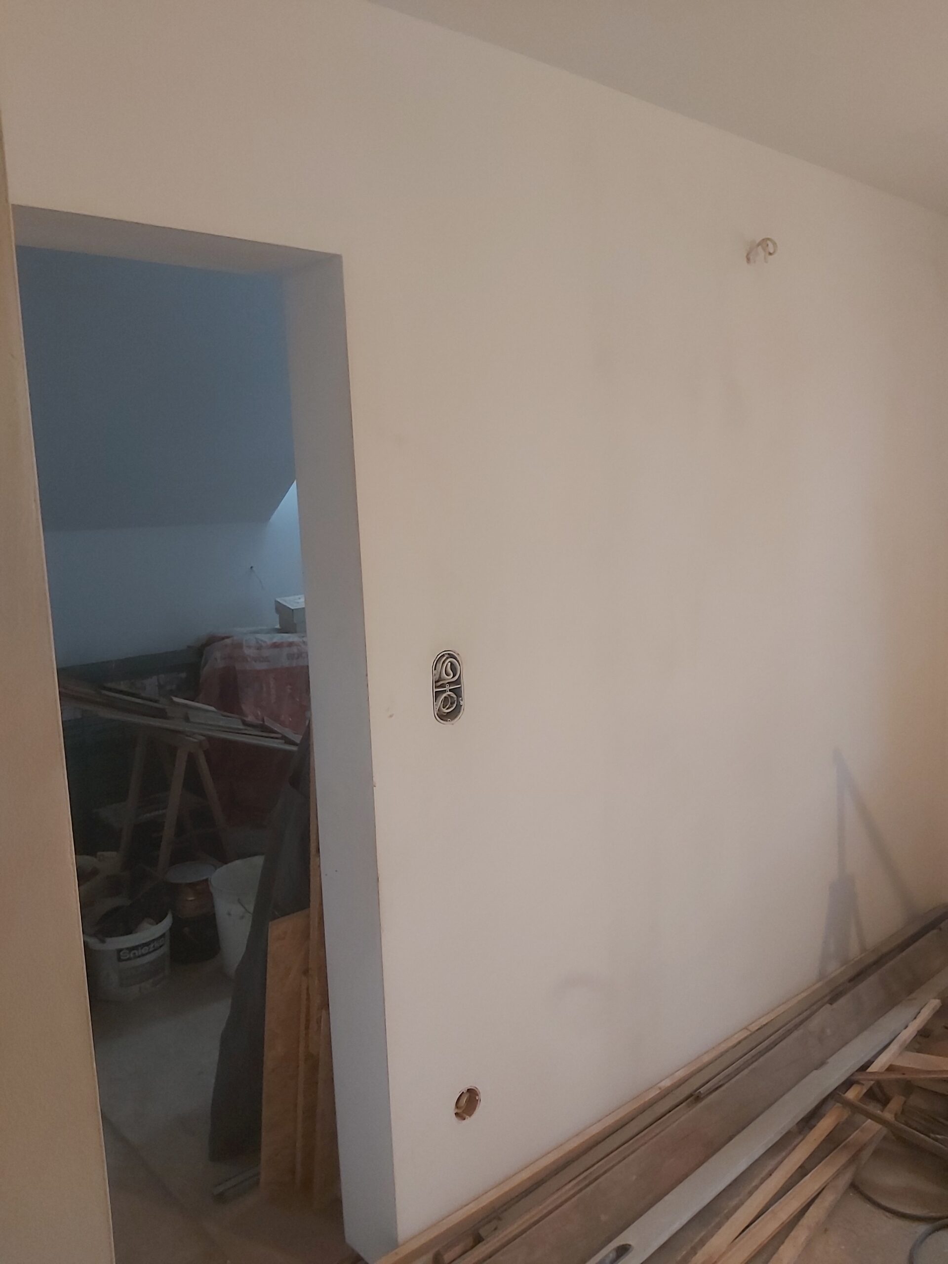 Wewnętrzna ściana po szpachlowaniu i malowaniu, z przygotowanym otworem drzwiowym oraz gniazdami elektrycznymi, w trakcie prac wykończeniowych.