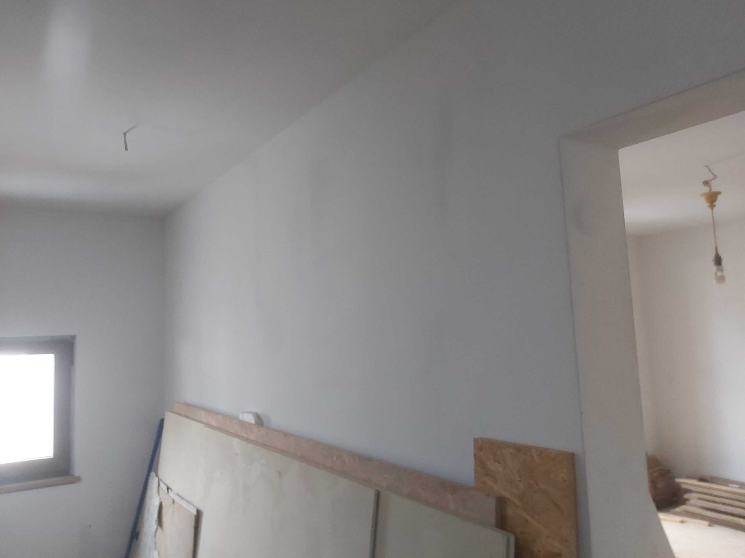 Jasna ściana wewnętrzna po wstępnym szpachlowaniu, przygotowana do dalszych prac wykończeniowych, z widocznym otworem drzwiowym i oknem.