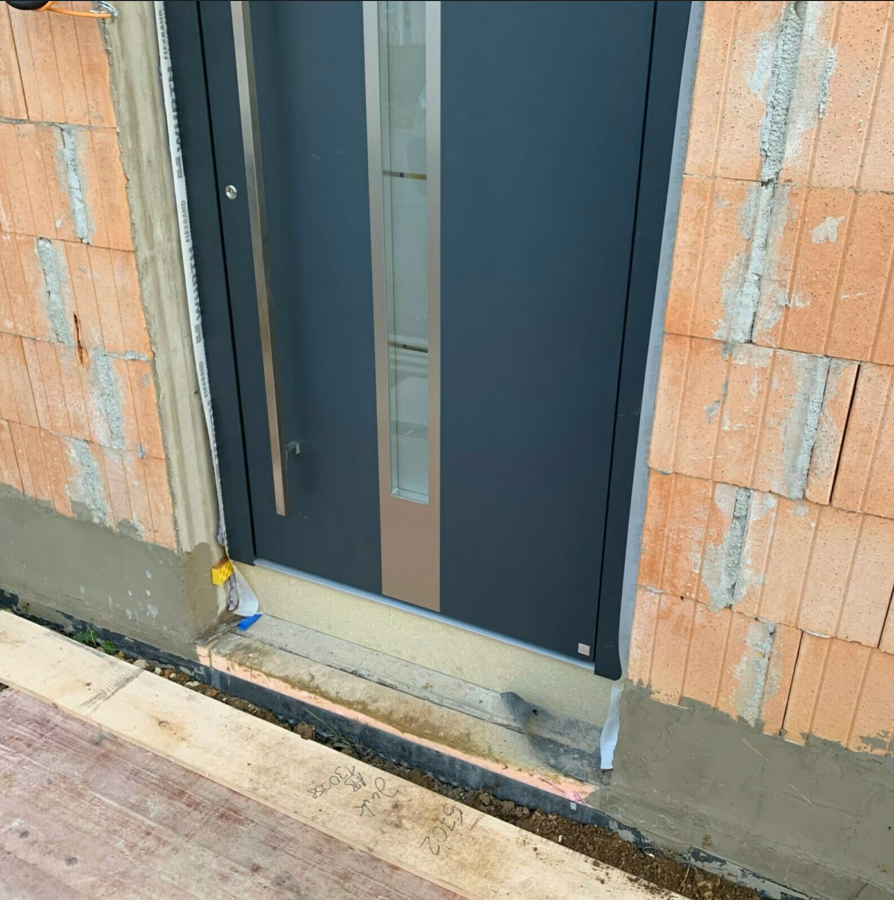 Zbliżenie na nowoczesne drzwi zewnętrzne w kolorze antracytowym, widoczny próg oraz dolna część ościeżnicy osadzone w murze z pustaków ceramicznych podczas montażu.