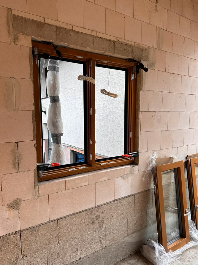 Dwuskrzydłowe okno z brązową ramą zamontowane w ścianie z bloczków, z widocznymi elementami montażowymi, przewodami i nieotynkowanymi ościeżami; obok oparte dodatkowe skrzydło okienne.