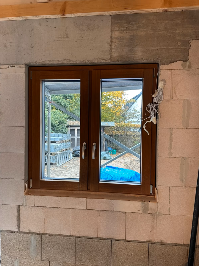 Dwuskrzydłowe okno z brązową ramą zamontowane w nieotynkowanym murze z bloczków, z widocznymi przewodami instalacyjnymi i surowym nadprożem betonowym.