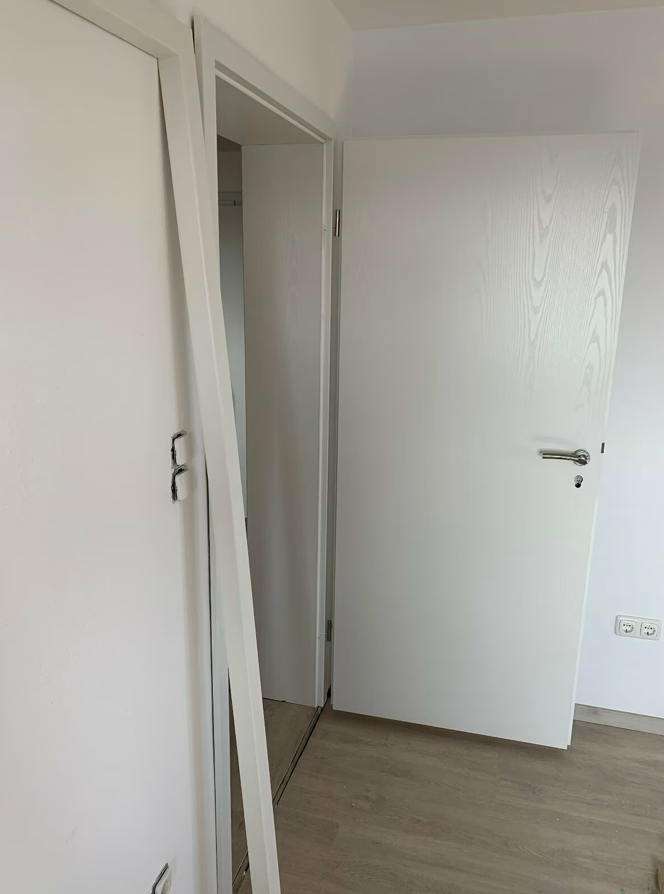 Białe drzwi wewnętrzne uchylone przy ościeżnicy, widok na jasne pomieszczenie z podłogą drewnianą po pracach wykończeniowych.