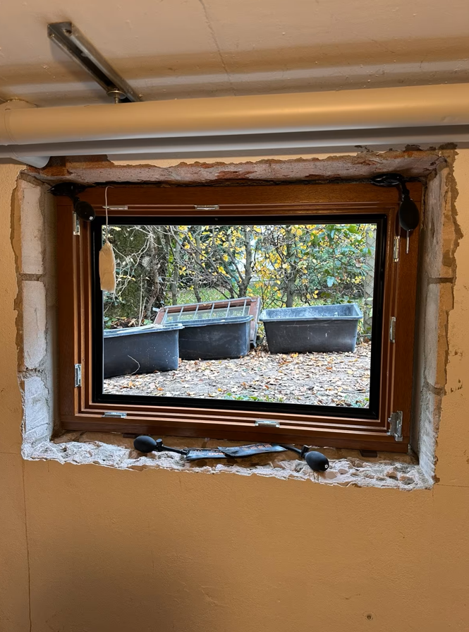 Okno piwniczne w brązowej ramie, zamontowane w surowym otworze okiennym bez obróbki tynkarskiej, z widoczną pianką montażową i nieotynkowanymi krawędziami muru.