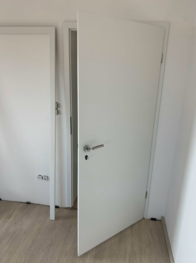 Białe drzwi wewnętrzne uchylone w jasnym pomieszczeniu, z metalową klamką i widoczną ościeżnicą po zakończonych pracach wykończeniowych.