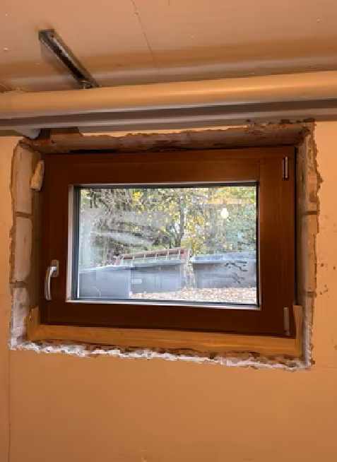 Małe okno piwniczne w brązowej ramie, osadzone w nieotynkowanym otworze okiennym, z widocznymi śladami montażu i surowymi krawędziami muru.