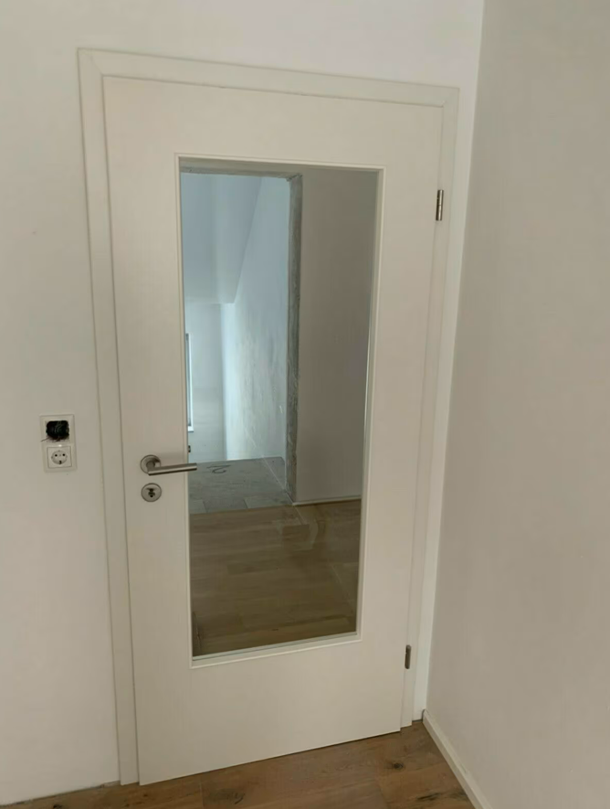 Białe drzwi wewnętrzne z pionowym przeszkleniem i metalową klamką, zamontowane w jasnym wnętrzu z drewnianą podłogą po remoncie.