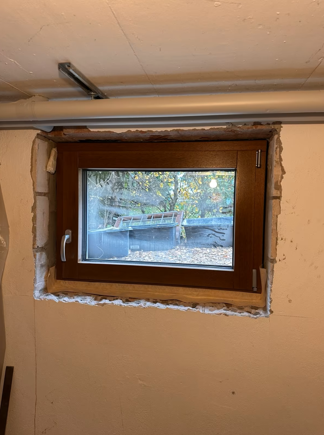 Okno piwniczne z brązową ramą osadzone w ścianie, widok od wewnątrz przed ostatecznym wykończeniem ościeża.