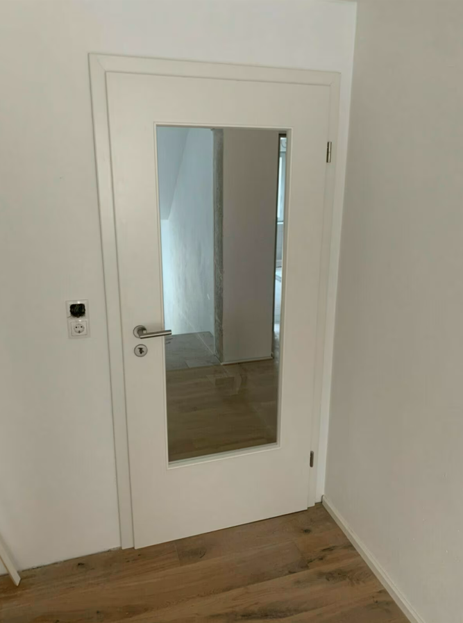 Białe drzwi wewnętrzne z pionowym przeszkleniem, zamontowane w jasnym wnętrzu z drewnianą podłogą po zakończonym remoncie.