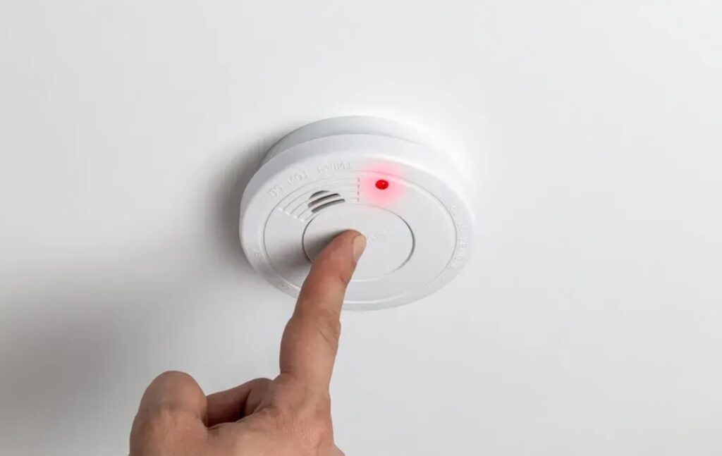 Ręka naciskająca przycisk testowy w sufitowym czujniku tlenku węgla z zapaloną czerwoną diodą sygnalizacyjną.