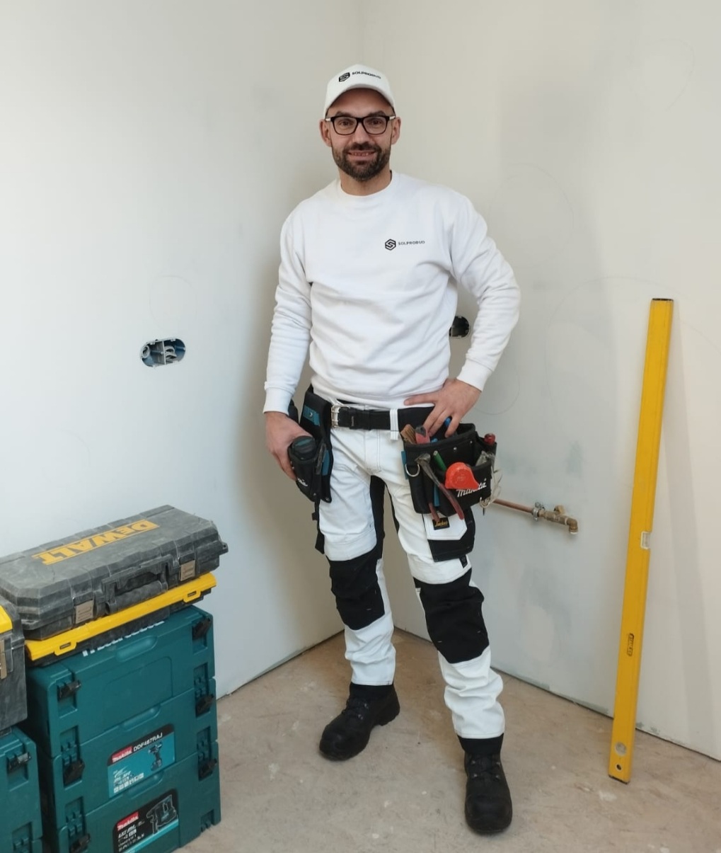 Instalator w odzieży roboczej z narzędziami przy pasie stoi w pomieszczeniu w trakcie prac montażowych, obok widoczne skrzynki narzędziowe i poziomica.