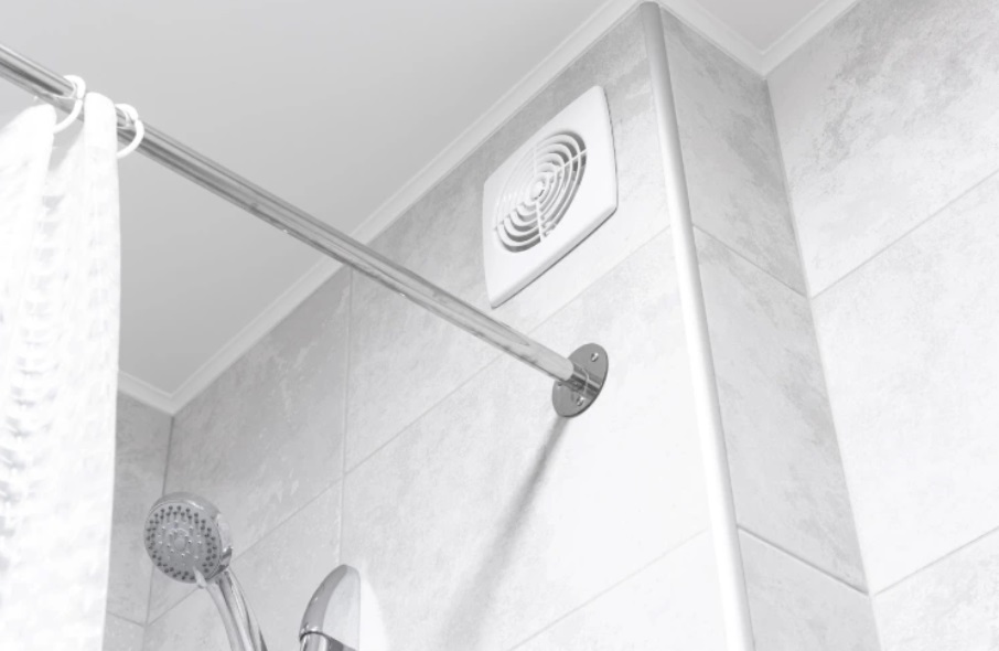 Biała kratka wentylacyjna zamontowana wysoko na ścianie łazienki nad kabiną prysznicową, obok drążka na zasłonę.