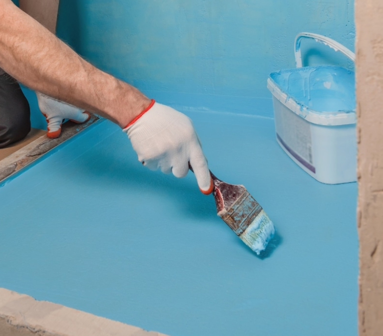 Zdjęcie przedstawia nakładanie niebieskiej folii w płynie pędzlem na podłogę i dolną część ściany w łazience, jako etap wykonywania hydroizolacji przed układaniem płytek.