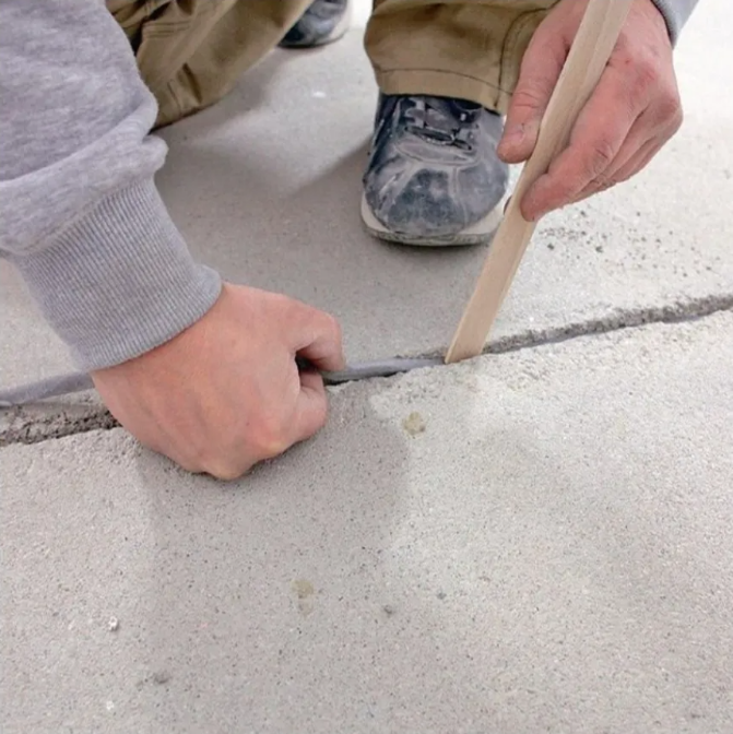 Zbliżenie na dłonie wykonawcy podczas wypełniania szczeliny dylatacyjnej w posadzce betonowej przed dalszym wykończeniem podłogi.