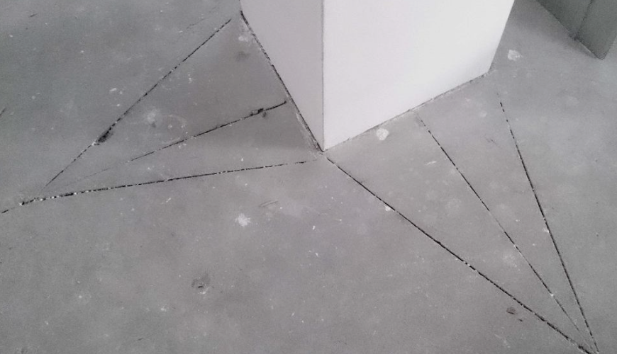 Widok posadzki betonowej z wykonanymi nacięciami dylatacyjnymi rozchodzącymi się od narożnika ściany, przygotowanymi do kontrolowania pracy podłoża i ograniczenia pęknięć.