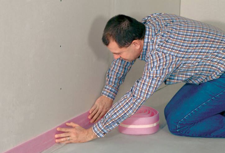 Mężczyzna montuje różową taśmę dylatacyjną przy ścianie na styku z posadzką, przygotowując podłoże do wykonania podłogi.