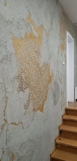 Ściana dekoracyjna przy drewnianych schodach, wykończona tynkiem strukturalnym z efektem postarzenia oraz ornamentowym wzorem w ciepłym odcieniu beżu.