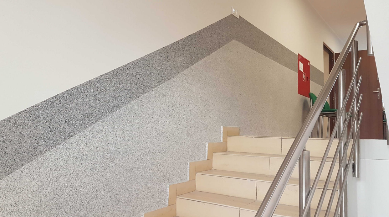 Klatka schodowa z balustradą metalową i dolną częścią ściany wykończoną szarym tynkiem mozaikowym, chroniącym powierzchnię przed zabrudzeniami i uszkodzeniami.